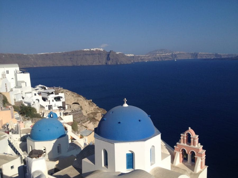 Dos iglesias con cúpulas azules, un campanario salmón, casas encaladas en lo alto de un acantilado en el pueblo de Oia, Santorini, y al fondo el mar Egeo azul oscuro y un largo acantilado