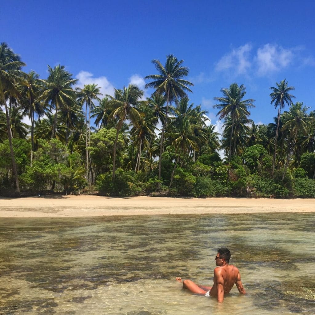 Pericles Rosa sentando en el agua rasa en la Playa de Moreré, Boipeba, Bahia, y algunos árboles ao fondo 