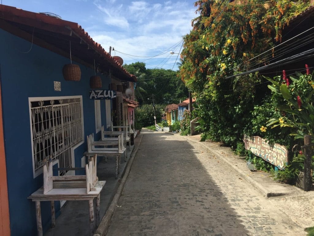 Una calle en el pueblo de Velha Boipeba, Bahia, Brasil, con una casa azul y ventanas blancas de un lado, y árboles en el otro lado