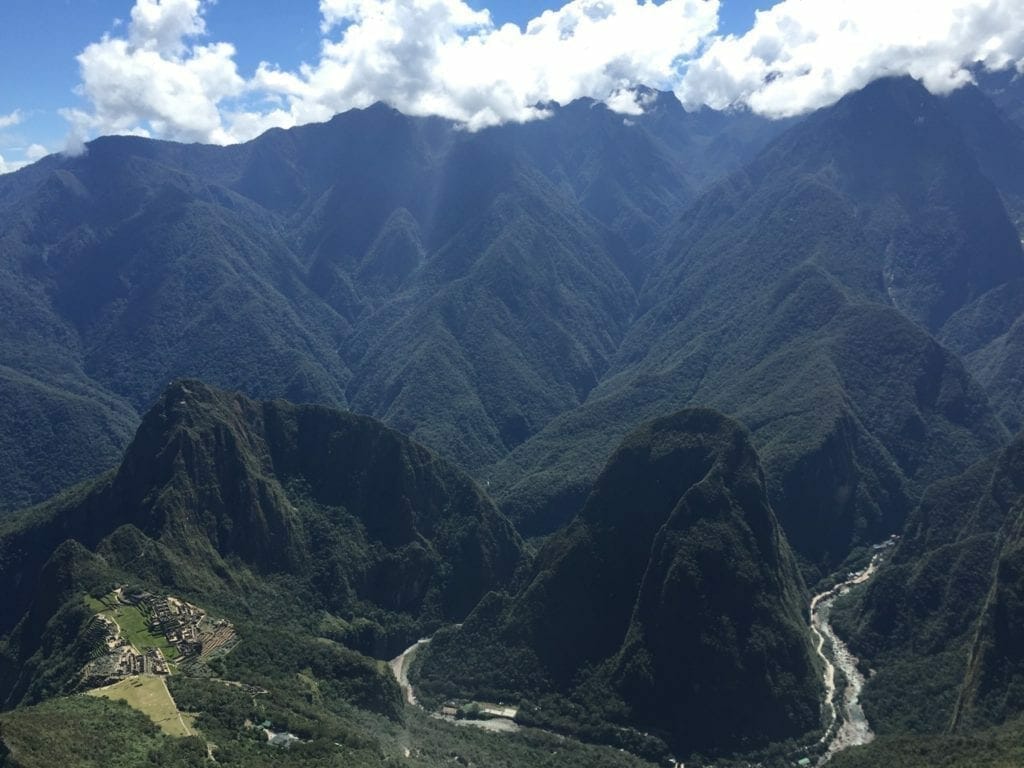 Um céu azul brilhante, nuvens e com rio Urubamba serpenteando ao redor das montanhas colossais cobertas com vegetação exuberante, Machu Picchu, Peru