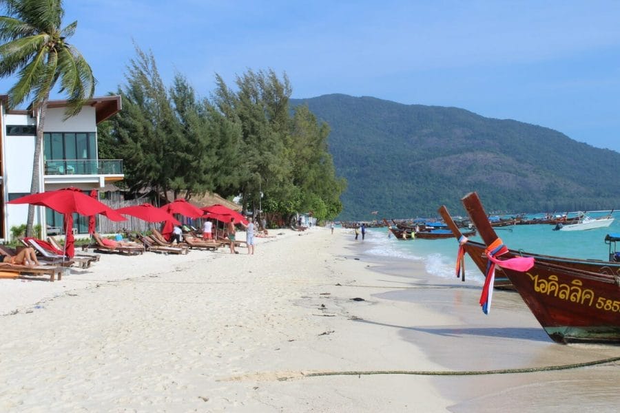 Sunrise Beach has white powdery sand, crystal-clear turquoise water, hotels, bars, some trees, long-tails boats on the water and a hill covered with luscious vegetation