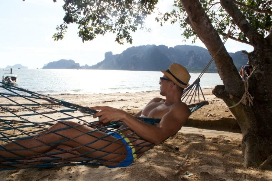 Homem de chapéu, óculos escuros e sunga azul em uma rede apreciando a bela vista de uma praia e morros cobertos de vegetação