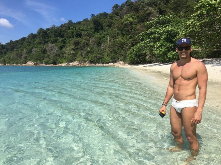 A deserted beach in the Tarutao National Marine Park with translucent water, white sand, a mountain covered with sumptuous vegetation and a man wearing a blue hat, sunglasses and a white speedo.