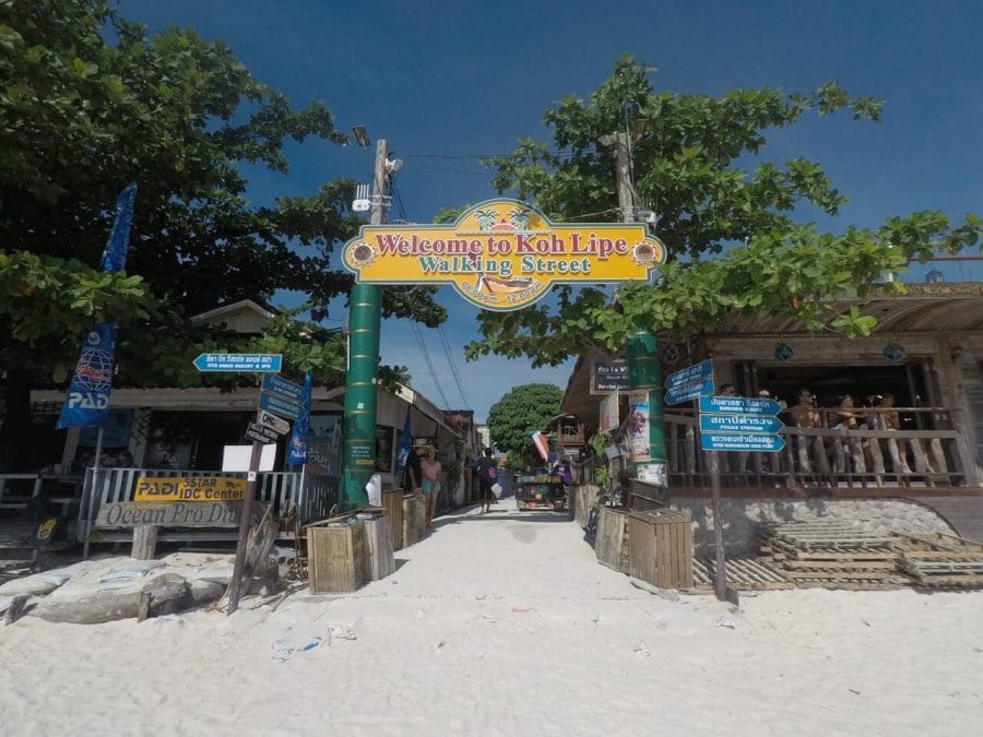 A rua principal da ilha de Ko Lipe, Tailândia, na entrada tem areai da praia, bares de madeira, árvores e um placa amarela onde se lê "Welcome to Koh Lipe - Walking Street"