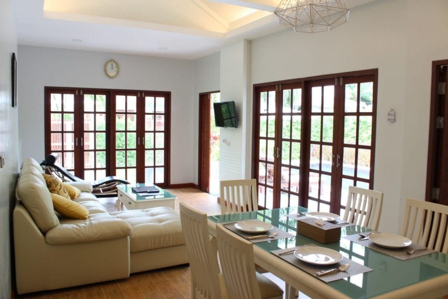 a sala de estar de uma villa do Alisea Pool Villas com mesa de jantar, cadeiras, sofá, almofadas, uma pequena tv na parede e portas de vidro com moldura de madeira