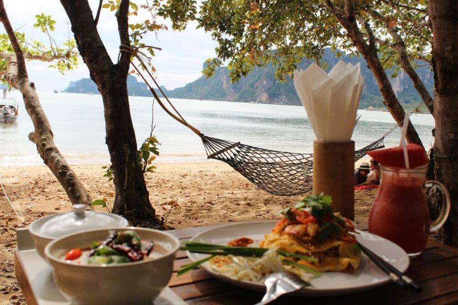 Mesa posta para o almoço no Adamana Beach Club, Aonang, com pratos, uma jarra de suco de melancia, uma rede e ao fundo o mar e as montanhas de calcário cobertas de vegetação