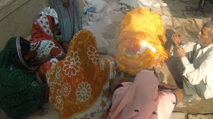 four women wearing traditional Indian saris and a man wearing a white shirt and a grey trouser squatting around a corpse covered with glittering golden cloths and flowers on a street in Varanasi
