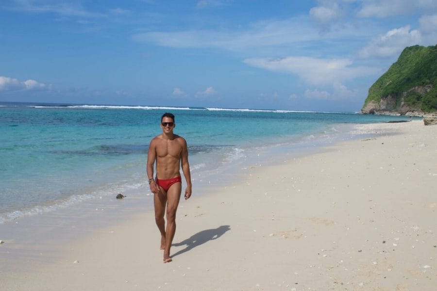 Pericles Rosa wearing a red swimsuit and sunglasses walking on Karma Beach, Bali, and the crystal-clear water and sea cliff covered with vegetation in the background