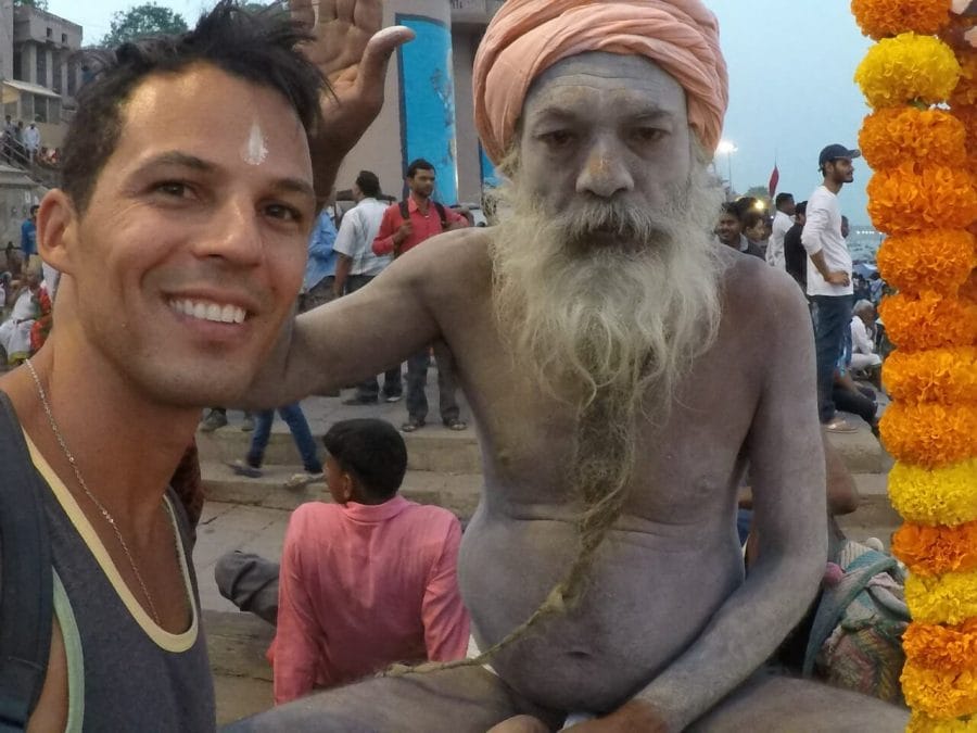 Pericles Rosa, from 7 Continents 1 Passport, wearing a grey tank top and a silver necklace with a Sadhu who has the body covered with ashes wearing a salmon colour turban on the Ganges River banks in Varanasi, India