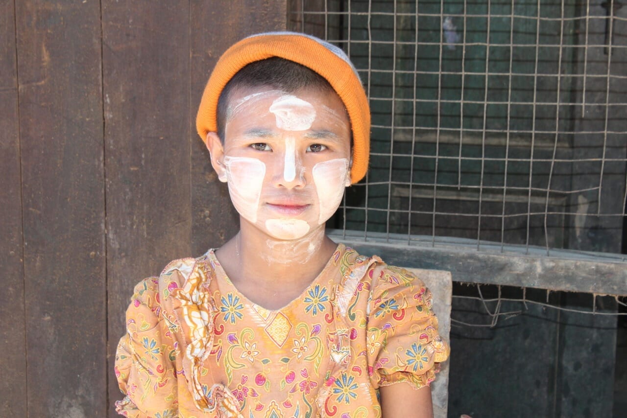 Um menino na frente da sua casa lendo um livro e usando thanaka, uma pasta cosmética branco-amarelada feita de casca de madeira usada frequentemente por locais.