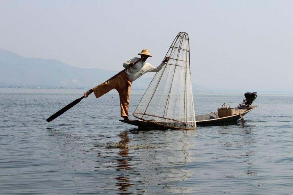 Um pescador remando com a perna e segurando um cesta de pescar em forma de cone no Inle Lake, Myanmar