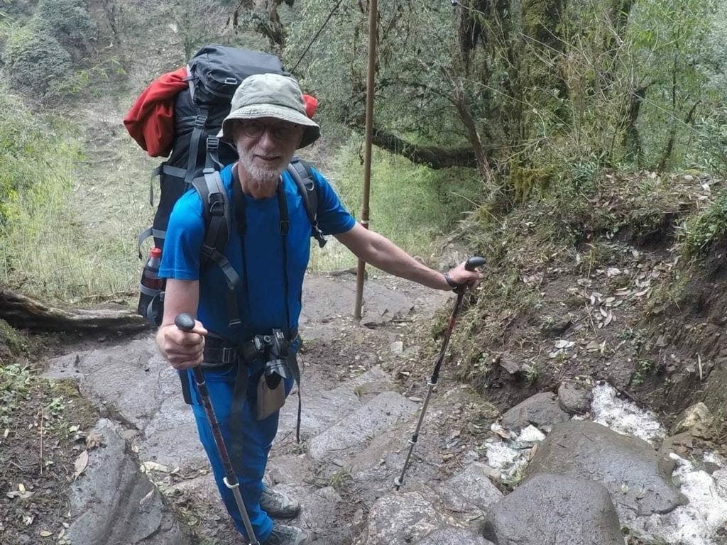 An elderly man wearing a blue t-shirt, blue trousers and olive green hat carrying a big backpack and holding two sticks climbing up some steps on a rhododendron forest in Ghorepani, Nepal