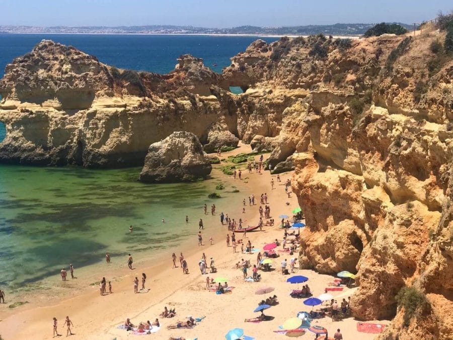 Pessoas passeando e tomando banho de sol na Prainha, Alvor, Portimão, Portugal, que é cercada por enormes falésias calcárias de cor ocre e tem águas verdes cristalinas.