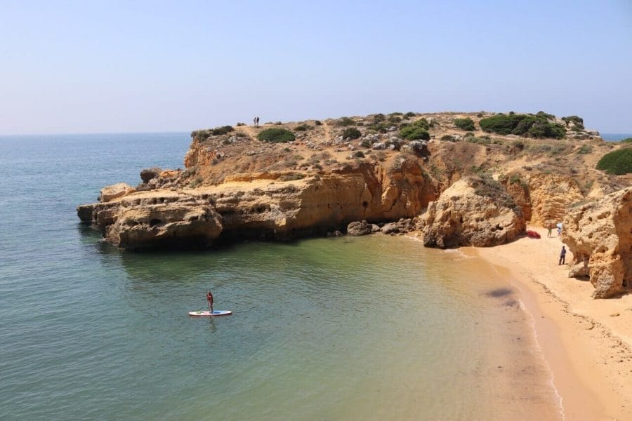 A person doing Standup paddleboarding on a section of Praia dos Arrifes, Albufeira, surrounded by yellow cliffs 