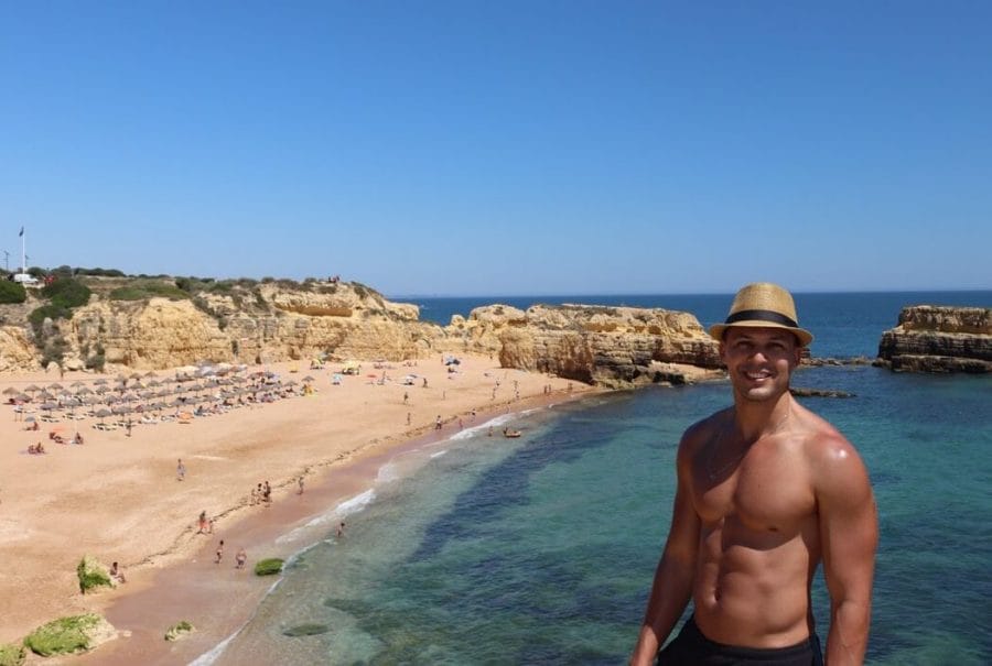 A man wearing a hat posing for a picture on the top a limestone cliff with Praia do Castelo in the background, Albufeira, Portugal