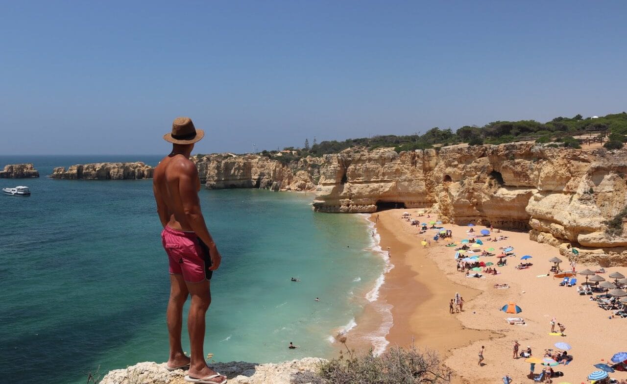 A man wearing a hat and red shorts on the top of a cliff overlooking the beach at Praia da Coelha, Albufeira, Algarve, Portugal