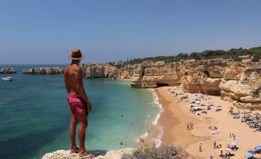 7 Most Beautiful & Best Beaches in Albufeira 1 A man wearing a hat and red shorts on the top of a cliff overlooking the beach at Praia da Coelha, Albufeira, Algarve, Portugal