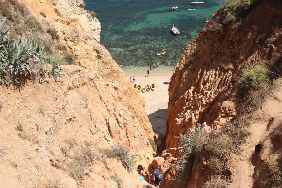 Playas de Lagos, Portugal: 7 Imperdibles + 1 Secreta 18 Acceso a Praia Grande con dos personas sosteniendo una cuerda entre acantilados anaranjados, kayaks y gente en la arena, botes y algunas personas y kayaks navegando por el agua.
