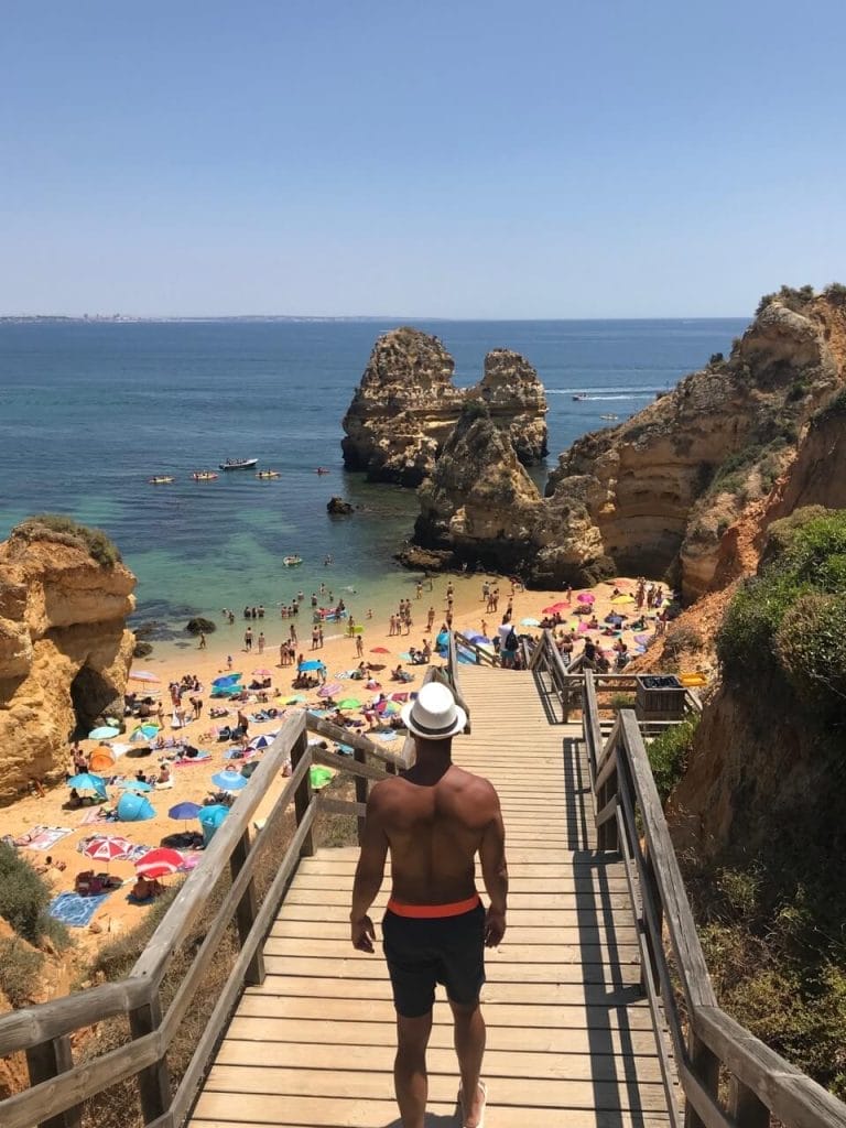 a man wearing a blue short and white hat standing on the wooden starway at Praia do Camilo in Lagos, that is surrounded by red-yellowish limestone cliffs and crystal-clear blue water, and some umbrellas and people on the beach