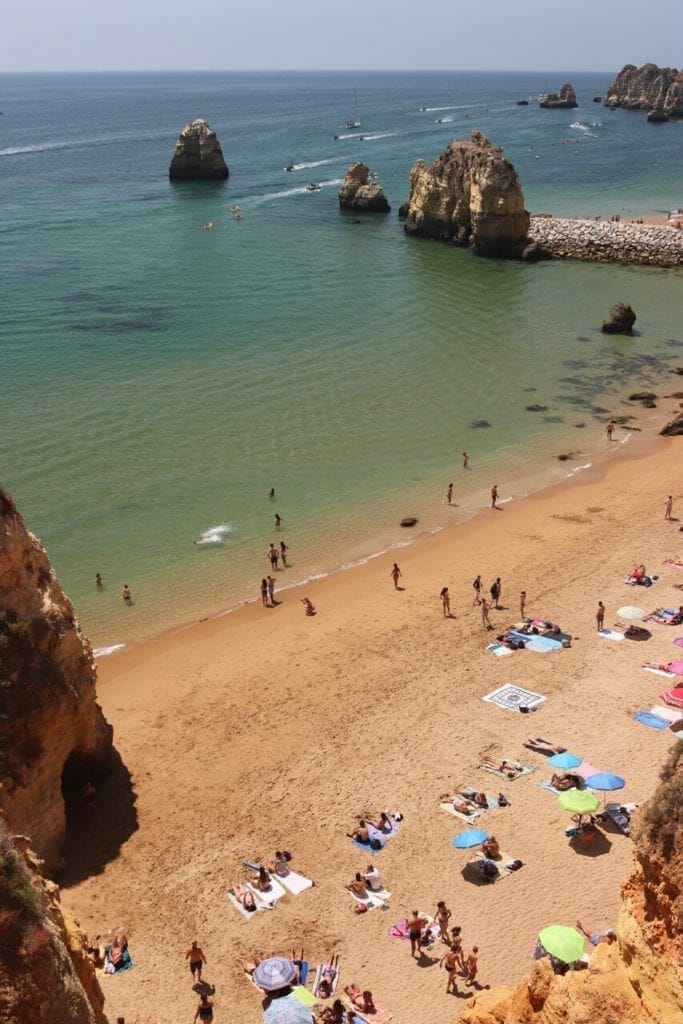 aerial view of Praia do Pinhão, with umbrellas of different clours, towels on the sand, people sunbathing and standing up, the crystal-clear water and sea cliffs