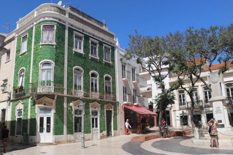 Uma praça na Cidade Velha de Lagos, Portugal, com casas caiadas de branco, um prédio de três andares coberto com azulejos verdes, algumas árvores no meio da praça e uma mulher tirando fotos de uma estátua