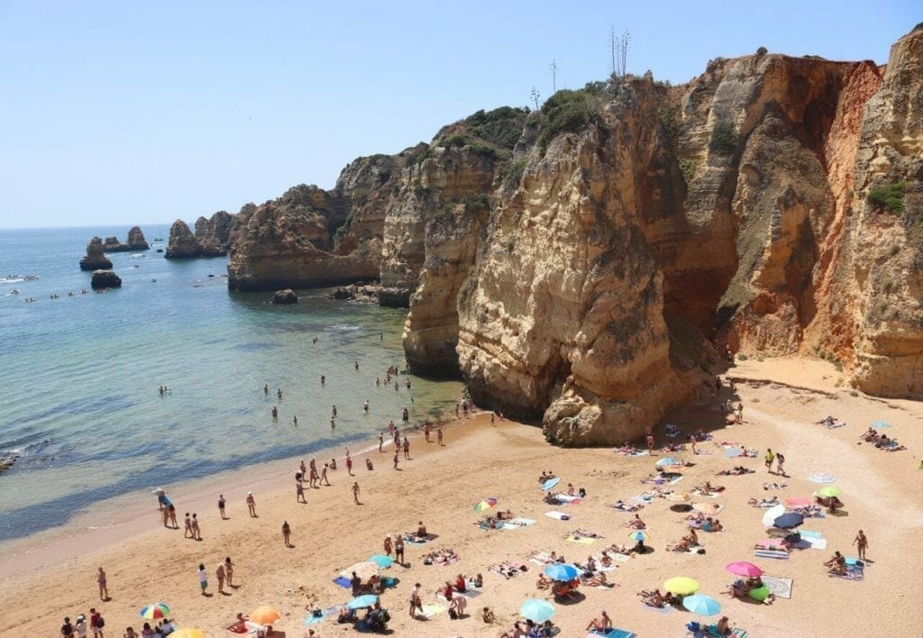 Praia da Dona Ana, Lagos, and its massive ochre limestone cliffs with people walking on the beach and sunbathing underneath umbrellas of different colours