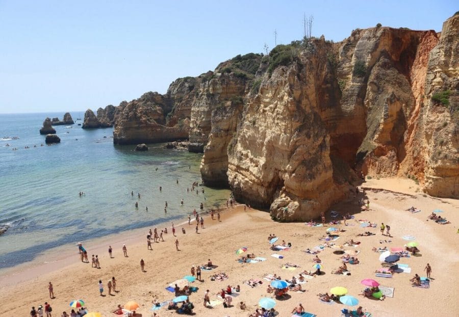 Praia da Dona Ana and its long and uneven wall, formed by ravishing orange cliffs covered in low vegetation, with people walking on the beach, sunbathing on towels and underneath umbrellas of different colours and others in the blue water.