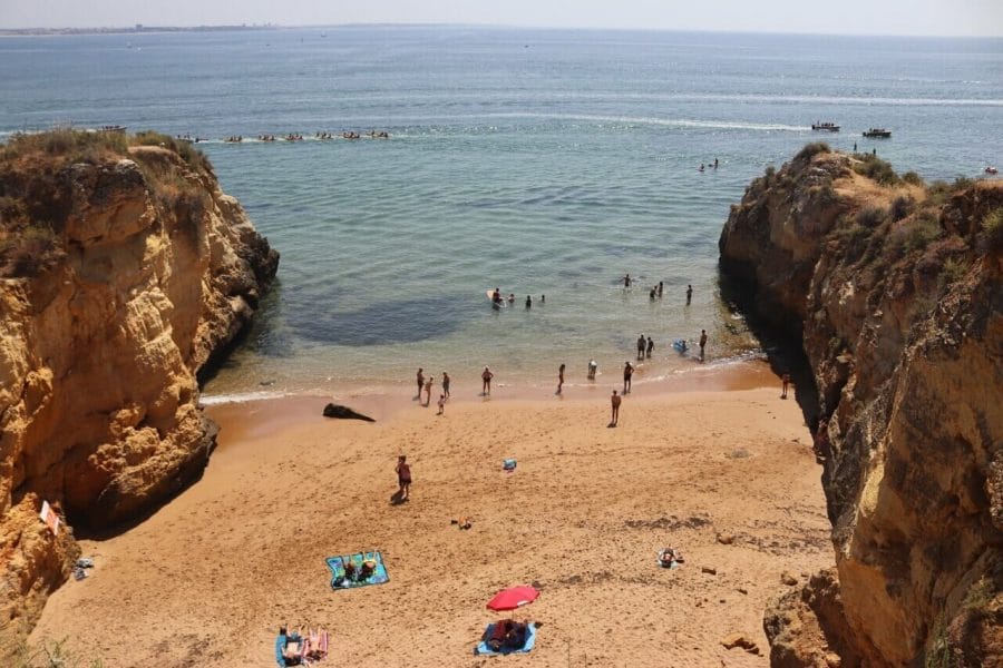 Playas de Lagos, Portugal: 7 Imperdibles + 1 Secreta 10 parte de Praia dos Estudantes, Lagos, con una playa rodeada de acantilados, gente tomando el sol sobre arena ocre y otras personas en el agua