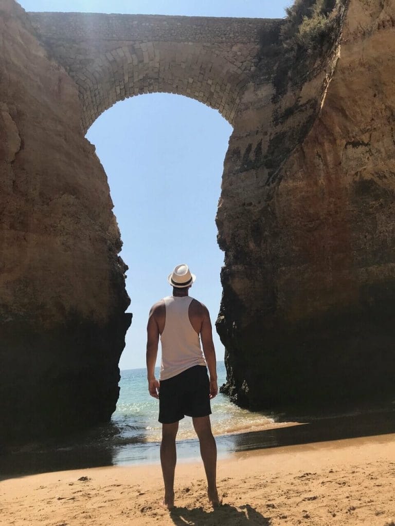Playas de Lagos, Portugal: 7 Imperdibles + 1 Secreta 12 un hombre vestido con pantalones cortos azules, una camiseta sin mangas blanca y un sombrero blanco frente a un arco en la Playa de los Estudiantes, Lagos, Portugal