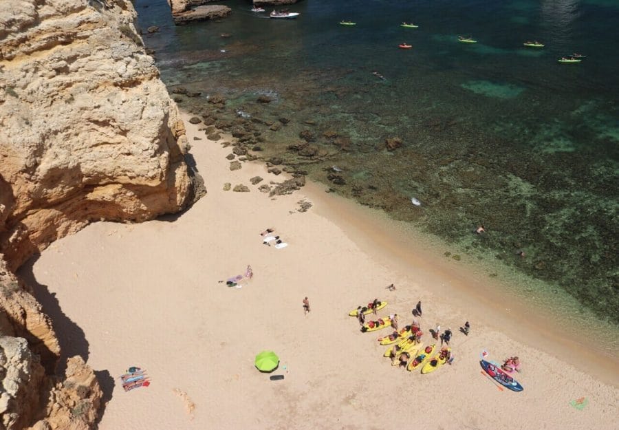 Playas de Lagos, Portugal: 7 Imperdibles + 1 Secreta 19 Vista aérea de Praia Grande con acantilado beige-naranja, gente acostada sobre toallas y en la arena cerca de kayaks amarillos, agua cristalina con algunos arrecifes de coral y kayaks verdes navegando en el agua