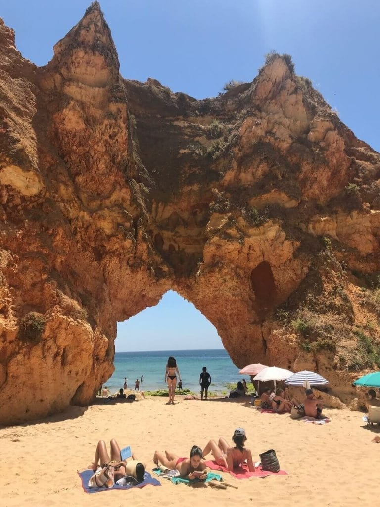 Três mulheres tomando banho de sol, quatro guarda-chuvas e uma moça passando por um majestoso arco na Praia dos Três Irmãos, Portimão, Portugal