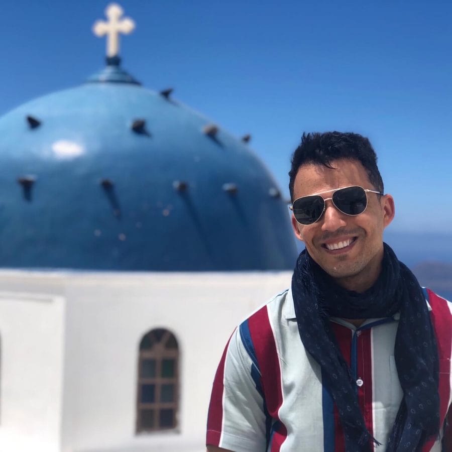 A man wearing sunglasses, dark blue scarf and a stripped shirt posing for a picture at Anastasi Church in the village of Imerovigli, Santorini
