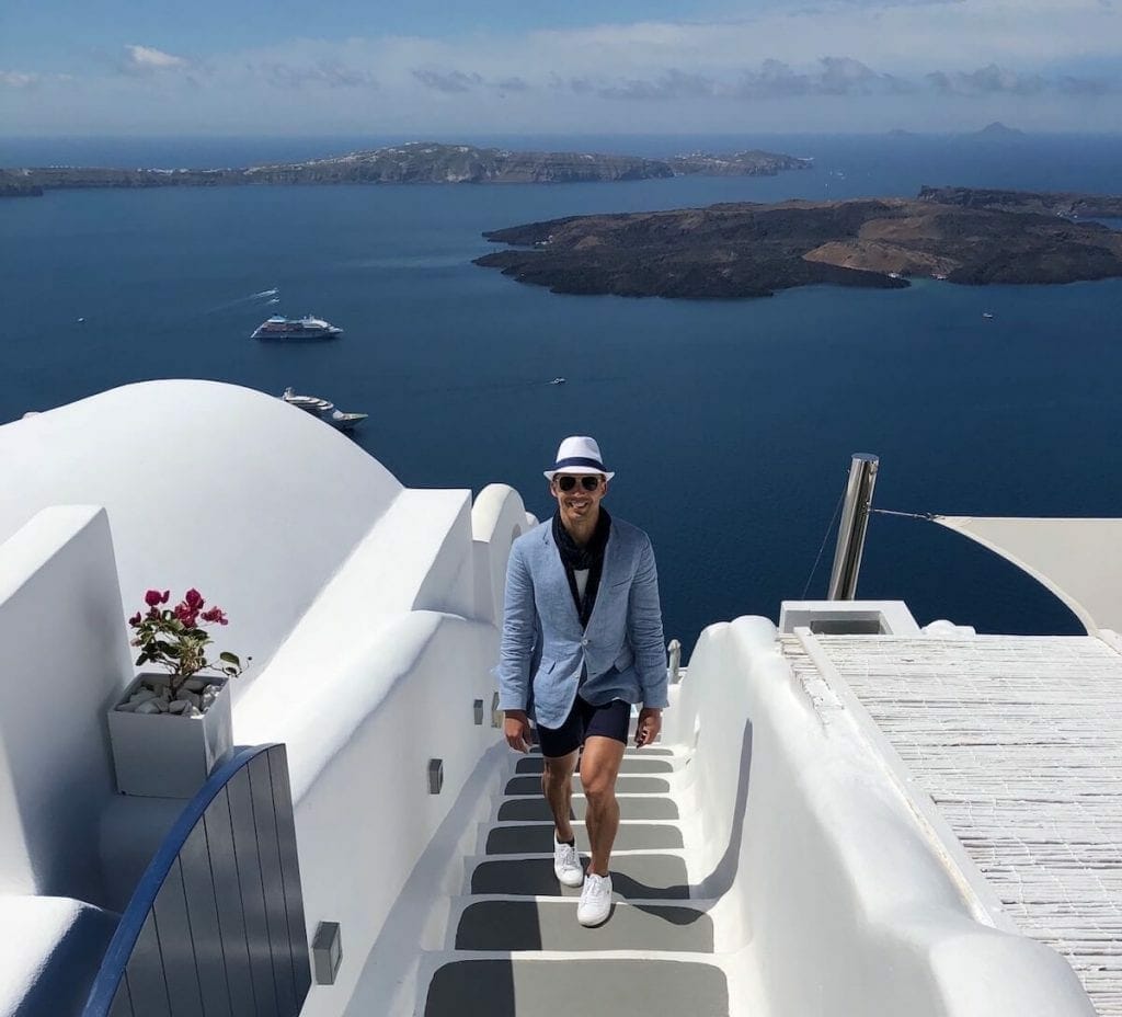 Pericles Rosa wearing a white hat, sunglasses, light blue blazer, marine blue scarf and shirt, white t-shirt and sneakers walking up the stairs of Chromata Hotel in Santorini.