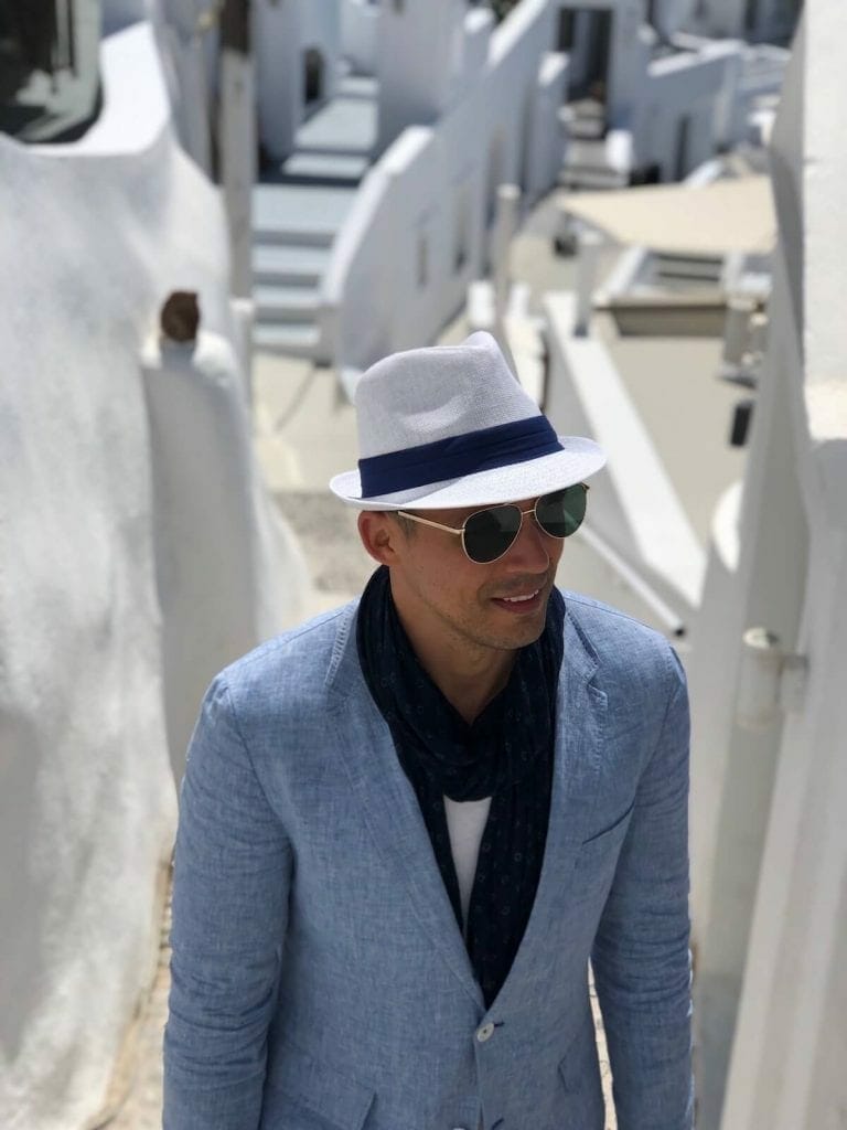 A man wearing a white hat, sunglasses, a light-blue blazer, a marine blue scarf, marine blue shorts and white trainers walking in an alleyway surrounded by whitewashed houses in the village of Imerovigli, Santorini, Greece