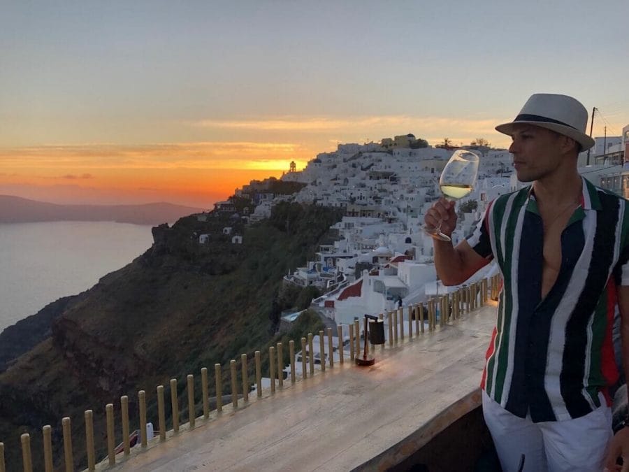 A man wearing a white hat and a stripped  shirt drinking a glass of white wine in the village of Imerovigli, Santorini, with the whitewashed houses in the background 