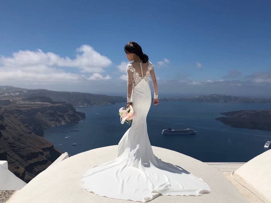 A bride posing for a picture on the top of a whitewashed house in Imerovigli, Santorini, with the Aegean Sea as a backdrop