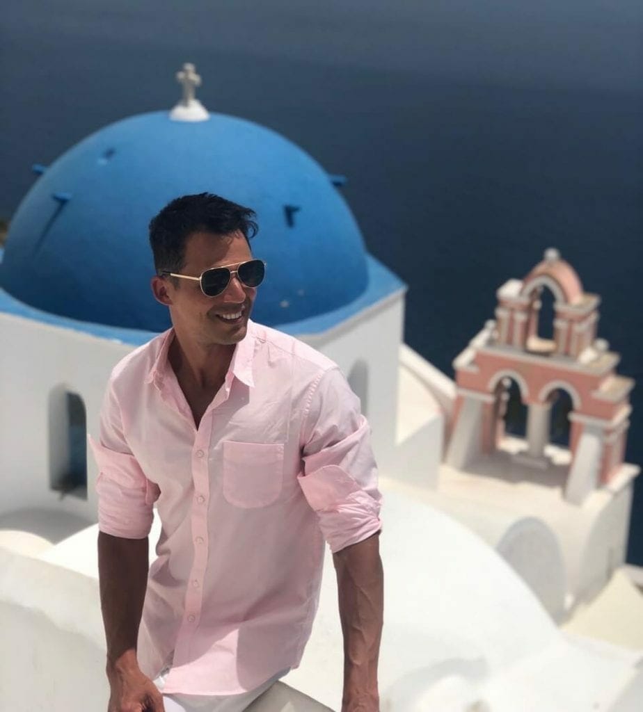 Pericles Rosa wearing sunglasses and a light pink shirt sitting on a white wall, a blue-domed church and a salmon bell tower behind him, in the village of Oia, Santorini