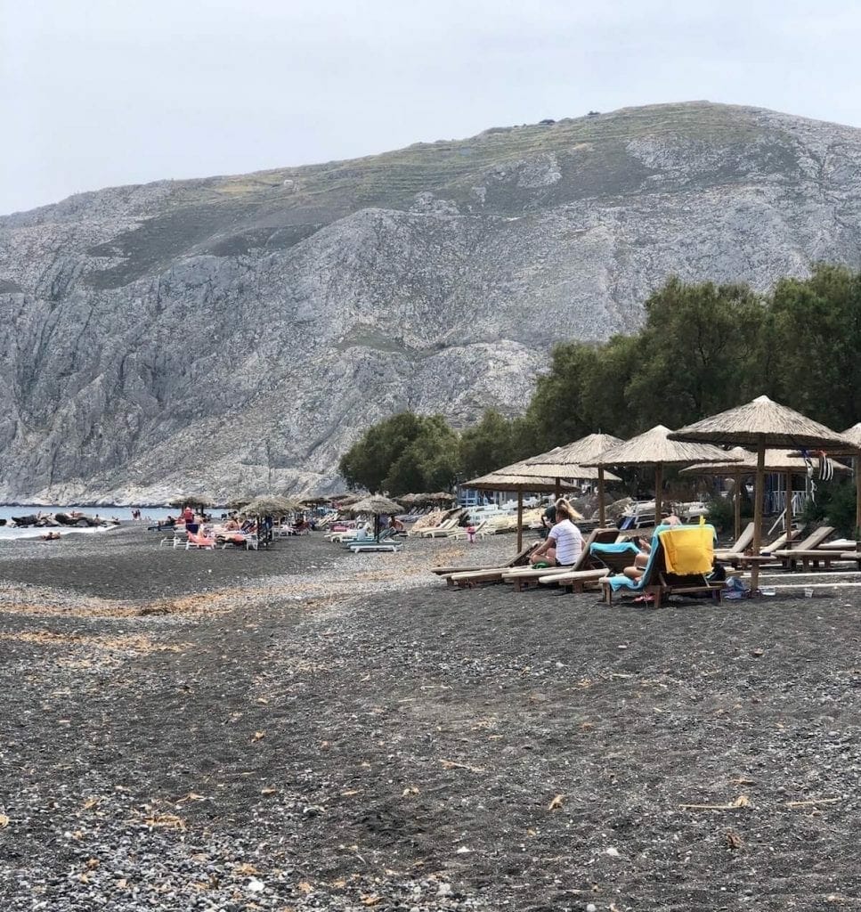 umbrellas, lounge chairs, a row of trees and a huge mountain in the background at Kamari Beach, Santorini