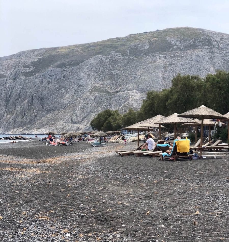 umbrellas, lounge chairs, a row of trees and a huge mountain in the background at Kamari Beach, Santorini