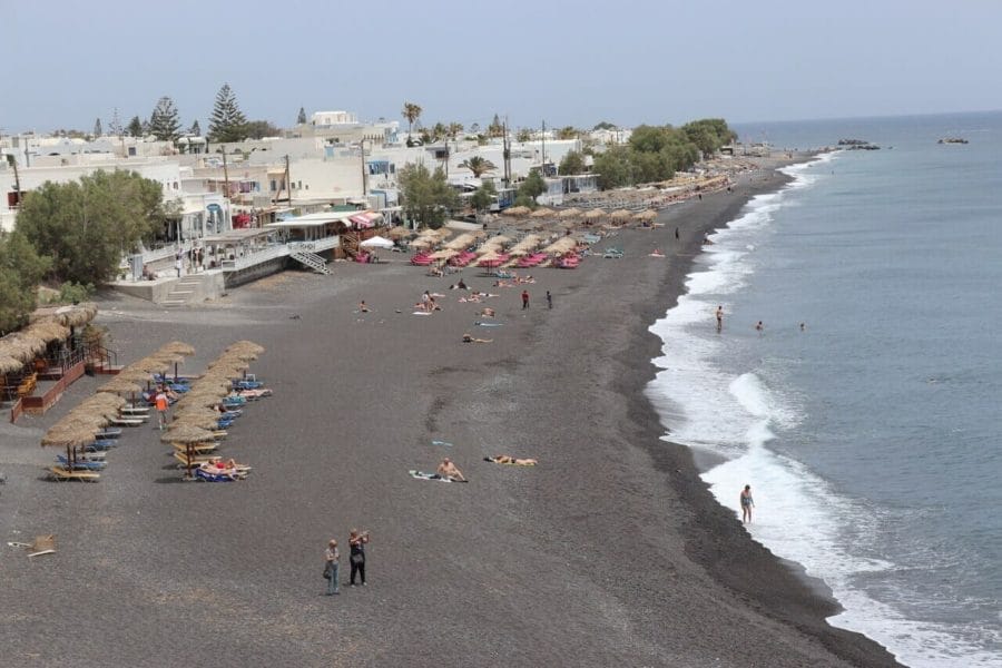 Some people walking on a black sand beach, umbrellas, sunbeds and whitewashed houses in the village of Kamari, Santorini
