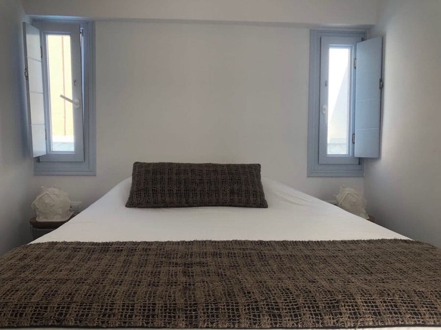 A room of San Giorgio hotel in Fira, Santorini, with a double bed with white sheets and brown pillow and tow small light-blue windows