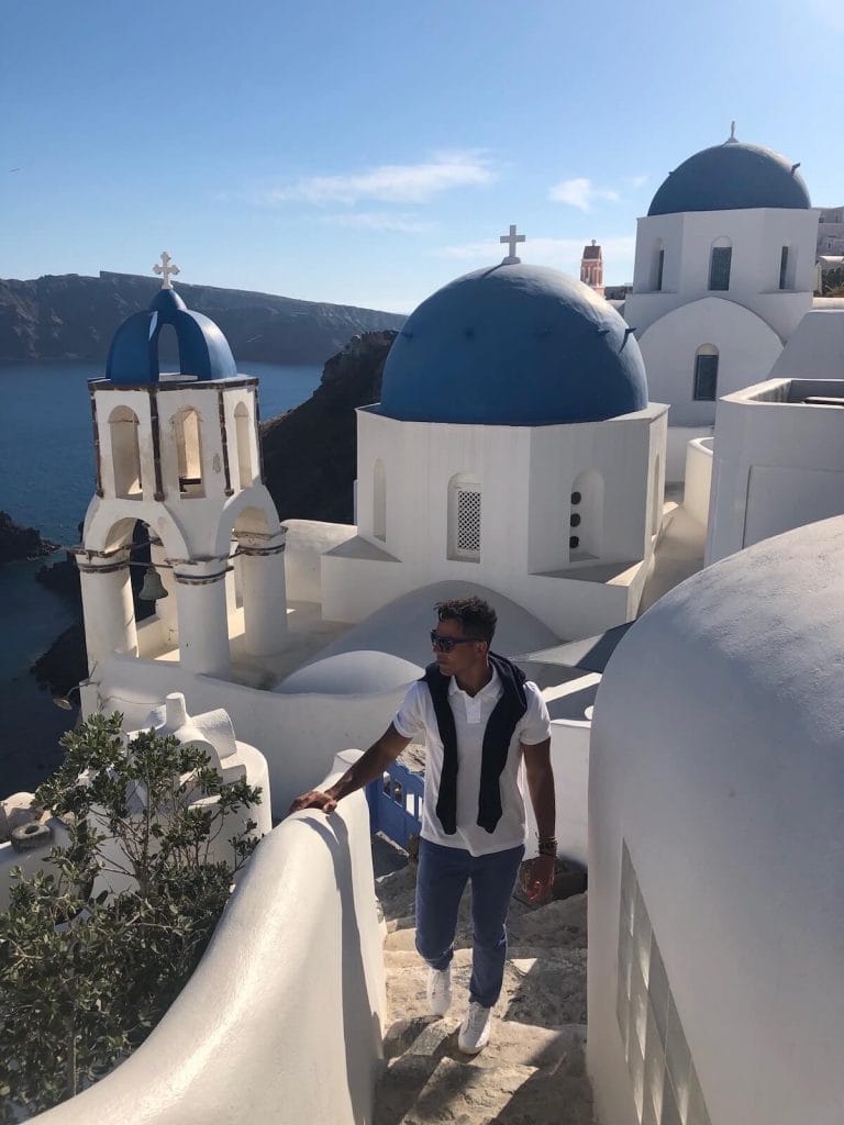 um homem de calça azul, tênis branco, polo branca, óculo de sol azul e suéter azul-marinho em rua uma estreita na vila de Oia, Santorini, e uma igreja branca com três cúpulas azuis ao fundo