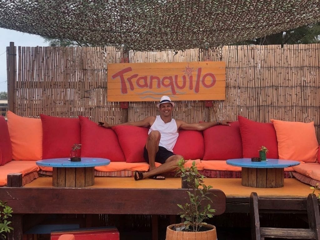 Pericles Rosa wearing a white hat, white tank top and blue shorts sitting on an orange couch with red pillows at Tranquilo Beach bar in Santorini
