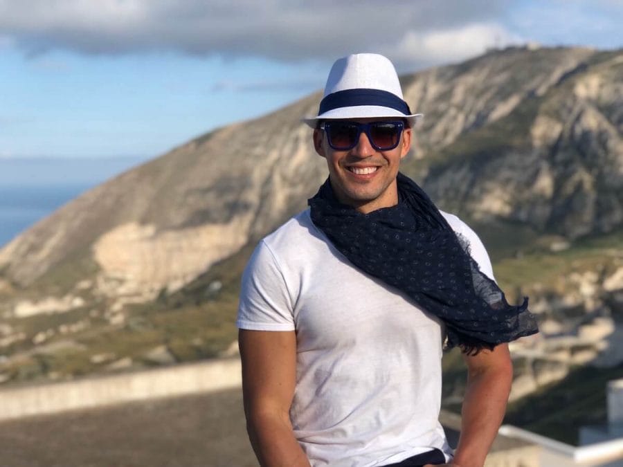 Pericles Rosa wearing a white hat, sunglasses, blue scarf and white t-shirt and a mountain in the background in the village of Pyrgos, Santorini