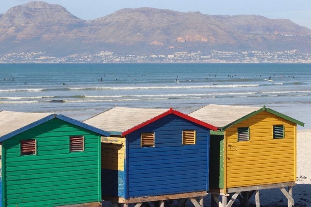 the colourful wood beach huts of Muizenberg Beach in Cape Town, South Africa, and in the background people surfing and a colossal mountain