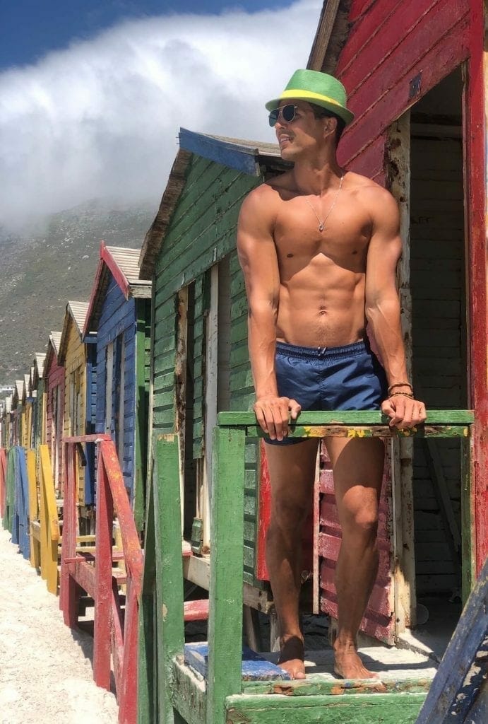 Pericles Rosa wearing a yellow and green hat, sunglasses and blue shorts in front of some colorful wooden beach huts in Muizenberg Beach, Cape Town, South Africa