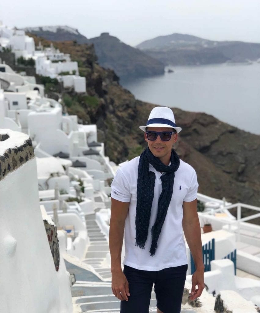 Un hombre con sombrero blanco, gafas de sol, bufanda azul marino, polo blanco y pantalones cortos azul marino en un callejón en Oia, Santorini, con algunas casas encaladas al fondo