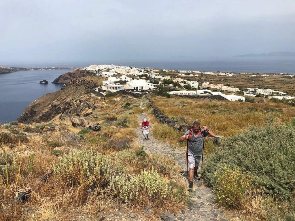 two people with hiking sticks walking the path between Fira and Oia and the village of Oia in the background