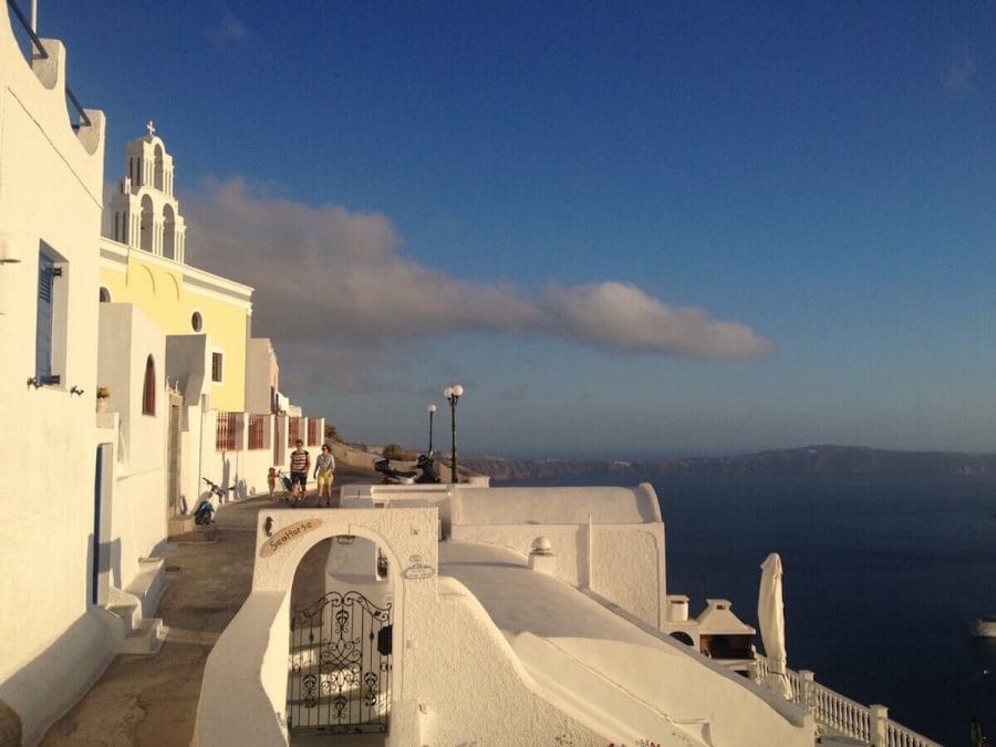 10 Best Things to Do in Fira 4 A couple walking by some whitewashed houses and an yellow church on the hiking trail from Fira to Oia and the Agean Sea in the background