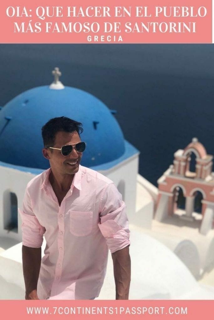 Un hombre con gafas de sol y una camisa rosa y una iglesia de cúpula azul con un campanario salmón y el mar Egeo al fondo, en la ciudad de Oia, Santorini